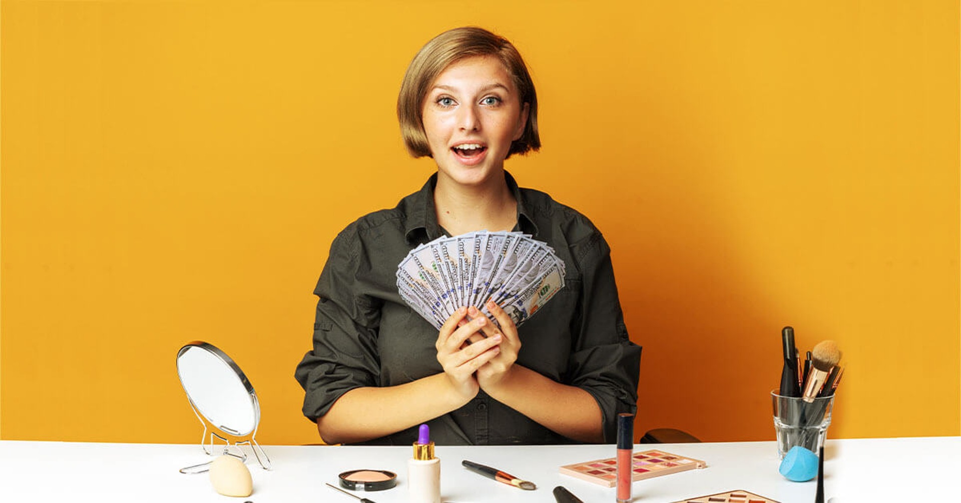 Female makeup vlogger holding hundred-dollar bills seated behind a table with small round mirror and makeup kits