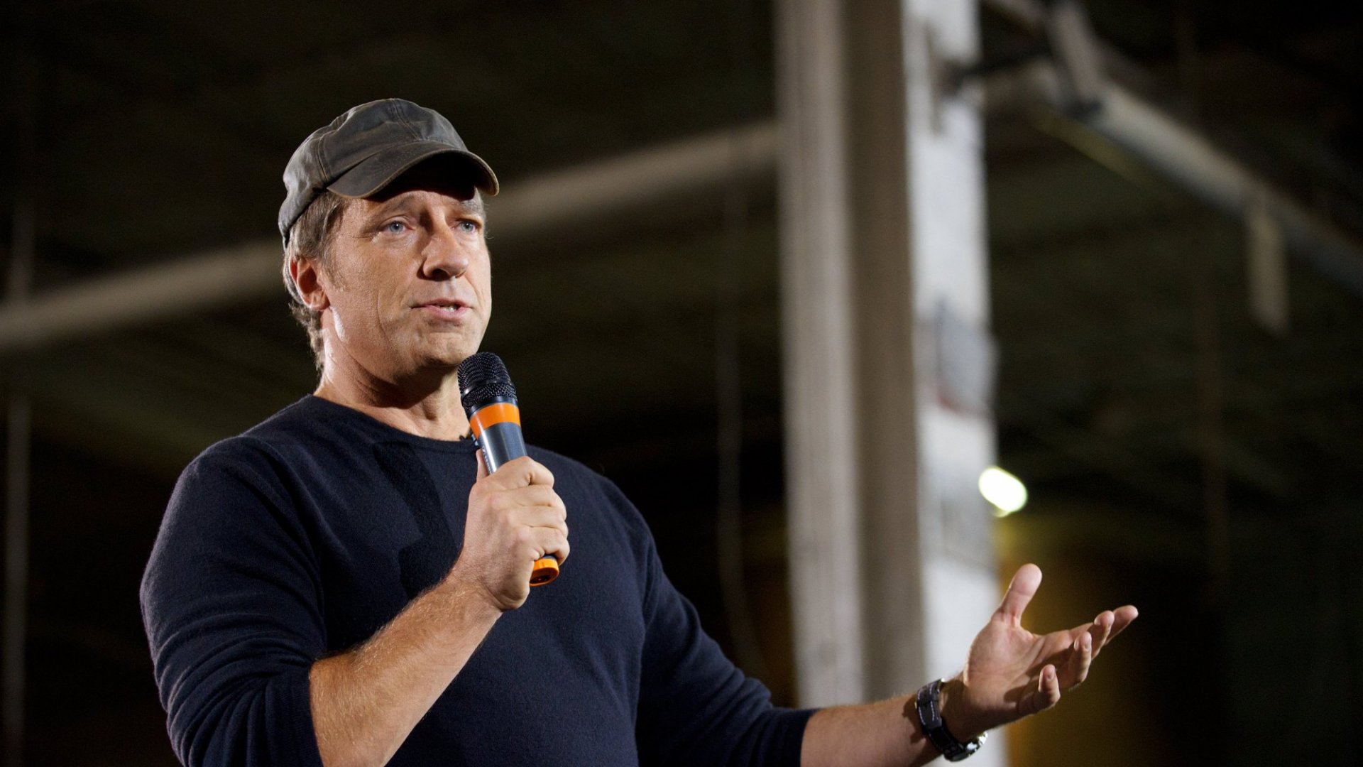 Mike Rowe wearing a black t-shirt and a face cap holing a mic while speaking