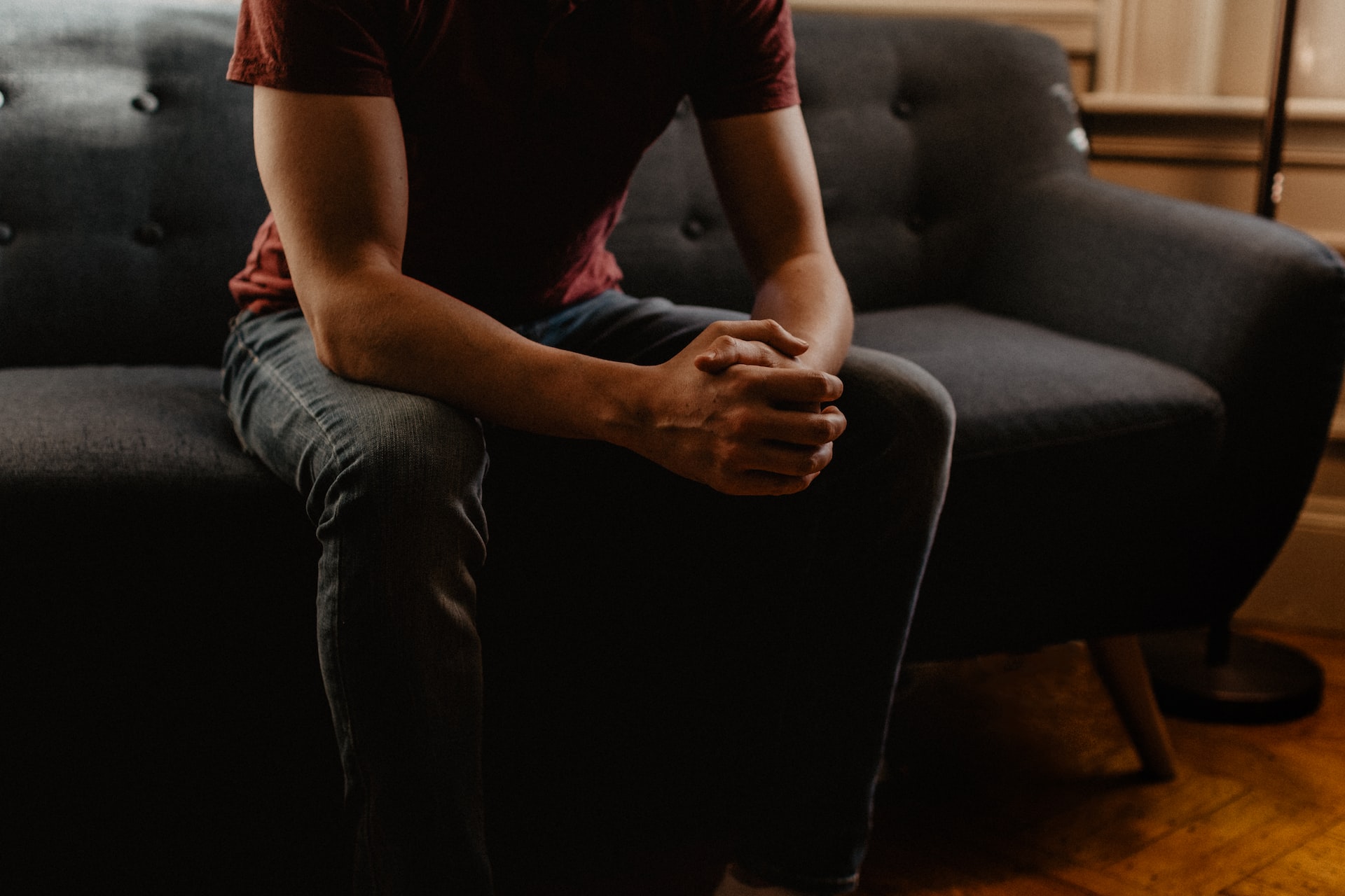 A guy on a sofa with hands clasped in plain maroon T-shirt and black denim pants having gay counseling