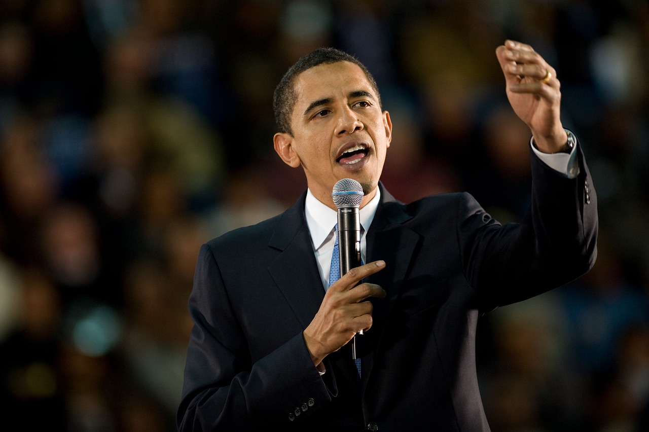 Barack Obama in a dark suit and light blue tie holding a wireless mic and gesturing while giving a speech