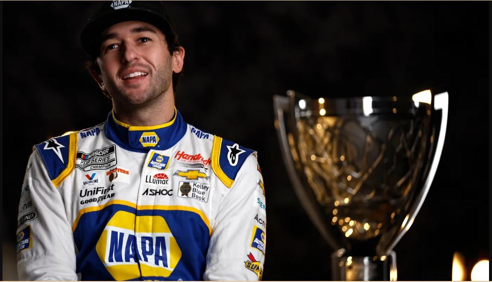 Chase Elliot wearing his racing attire while sitting close to his NASCAR trophy