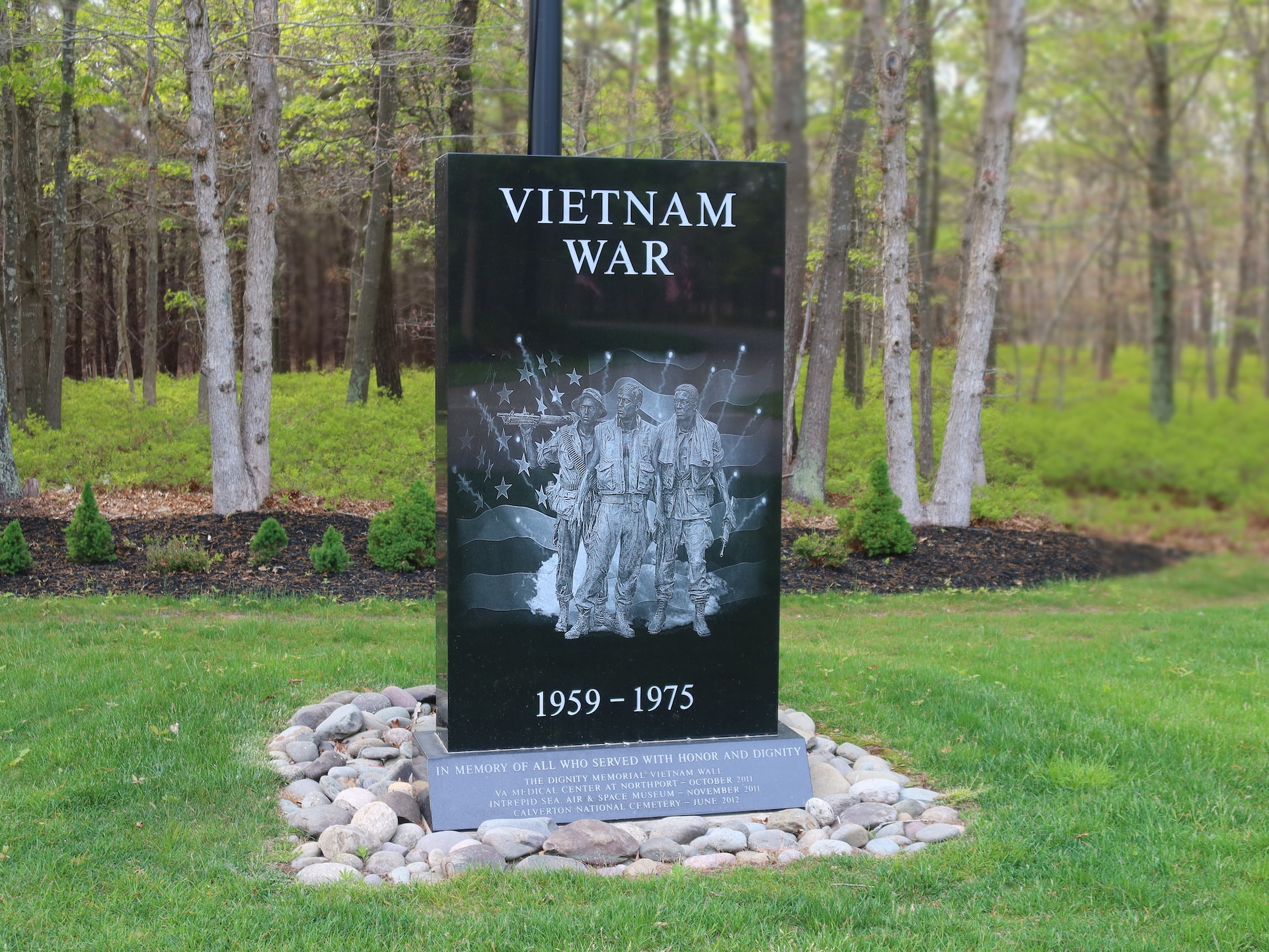 A Vietnam War memorial made of black polished stone set on a pile of small smooth rocks in a grassy wooden area