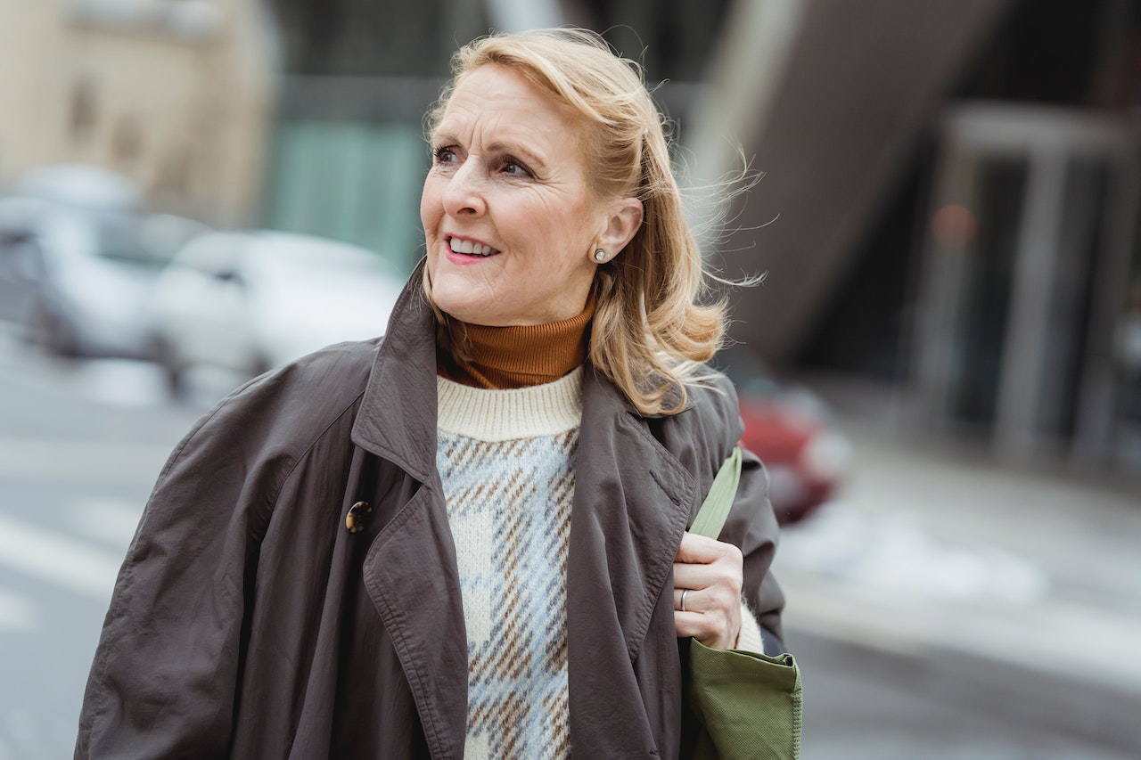 A smiling blonde woman with no gray hair at 65 on the street in coat and turtleneck shirt under a sweatshirt
