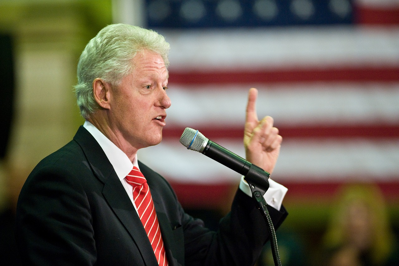 Bill Clinton in dark suit and red tie talking into a microphone and gesturing with his left hand