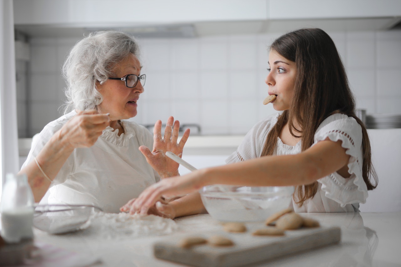 A grandmother with gray hair with granddaughter both in white clothes making cookies on the kitchen