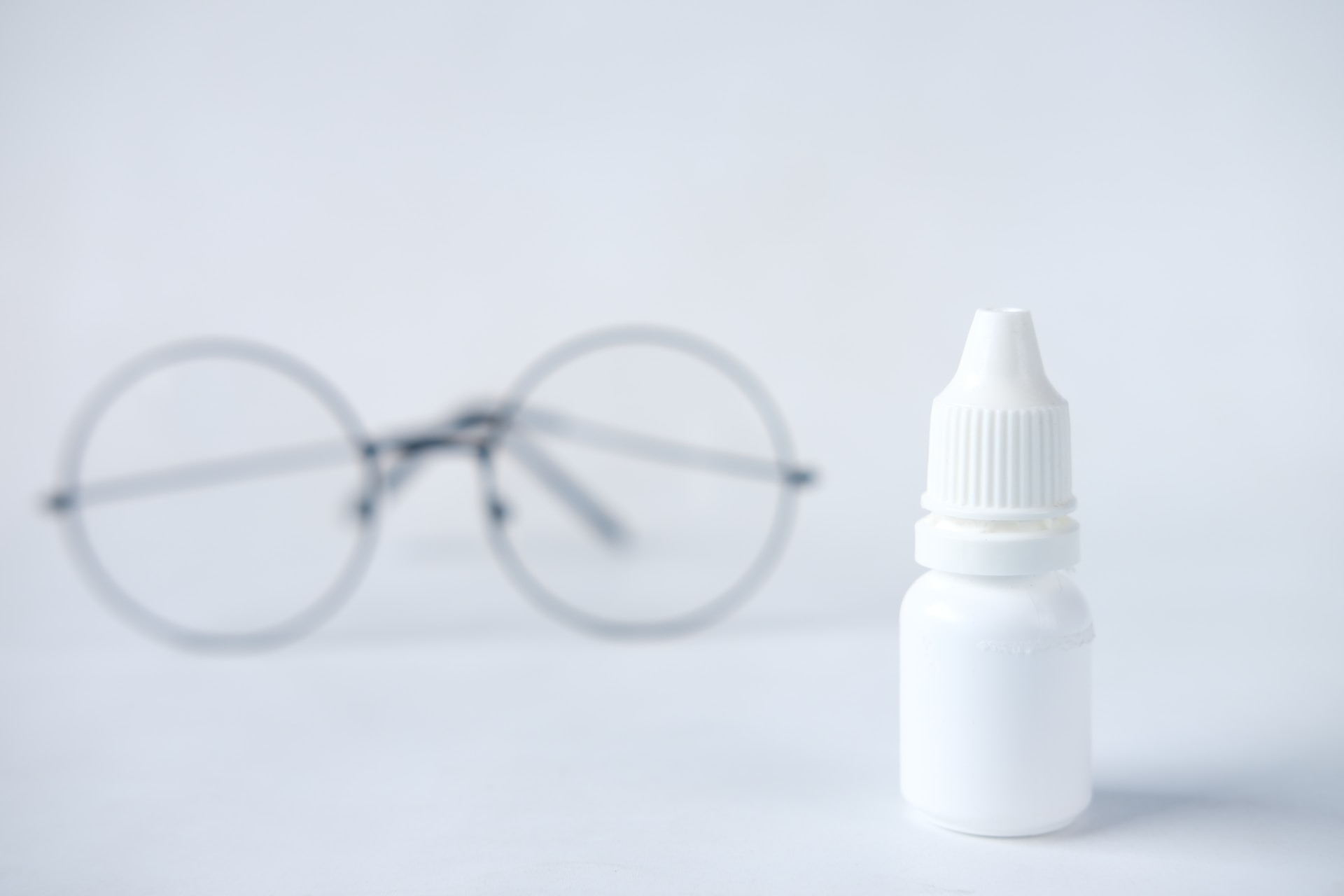 Small eye drops in unlabeled white container, with a black-rimmed round eye glasses blurred in the background