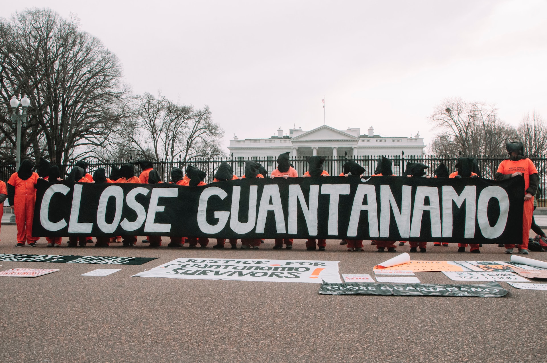 Protesters across the White House in orange clothes and heads covered hold a sign that says ‘Close Guantanamo’