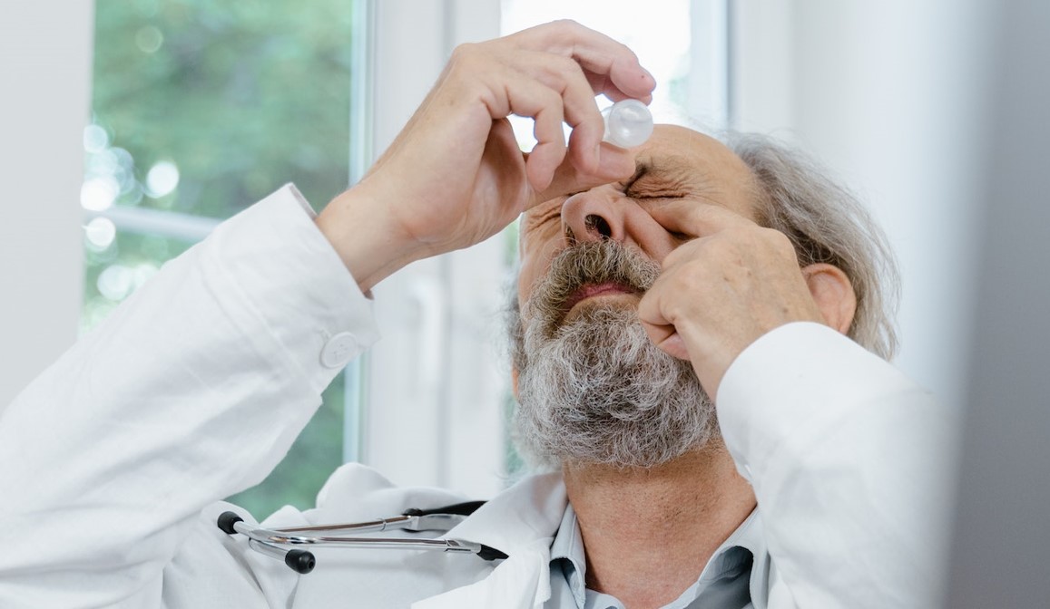 A gray-haired and bearded male eye doctor in his clinic using eye drops