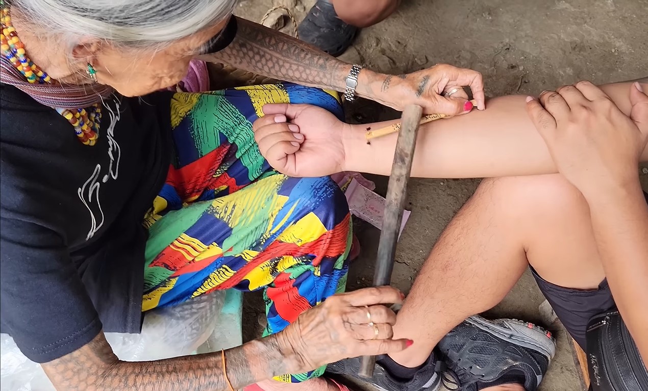 Apo Whang-od in dark blue T-shirt and colorful loose pants doing her three dots tattoo on a male’s pulse