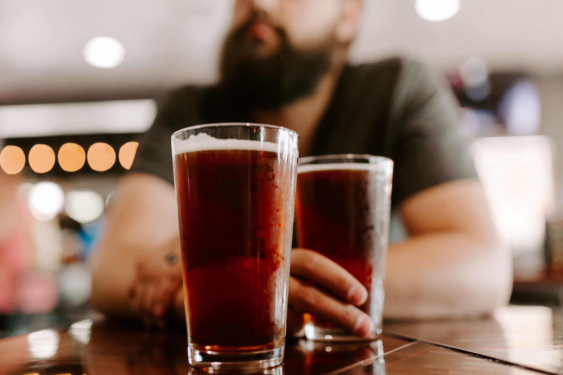 Two glasses of dealcoholized beer on a table, with a blurred bearded guy holding one of them