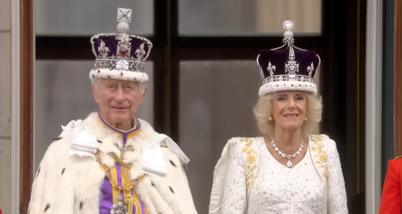 A smiling King Charles III and Queen Camilla with their crowns at the balcony of Buckingham Palace