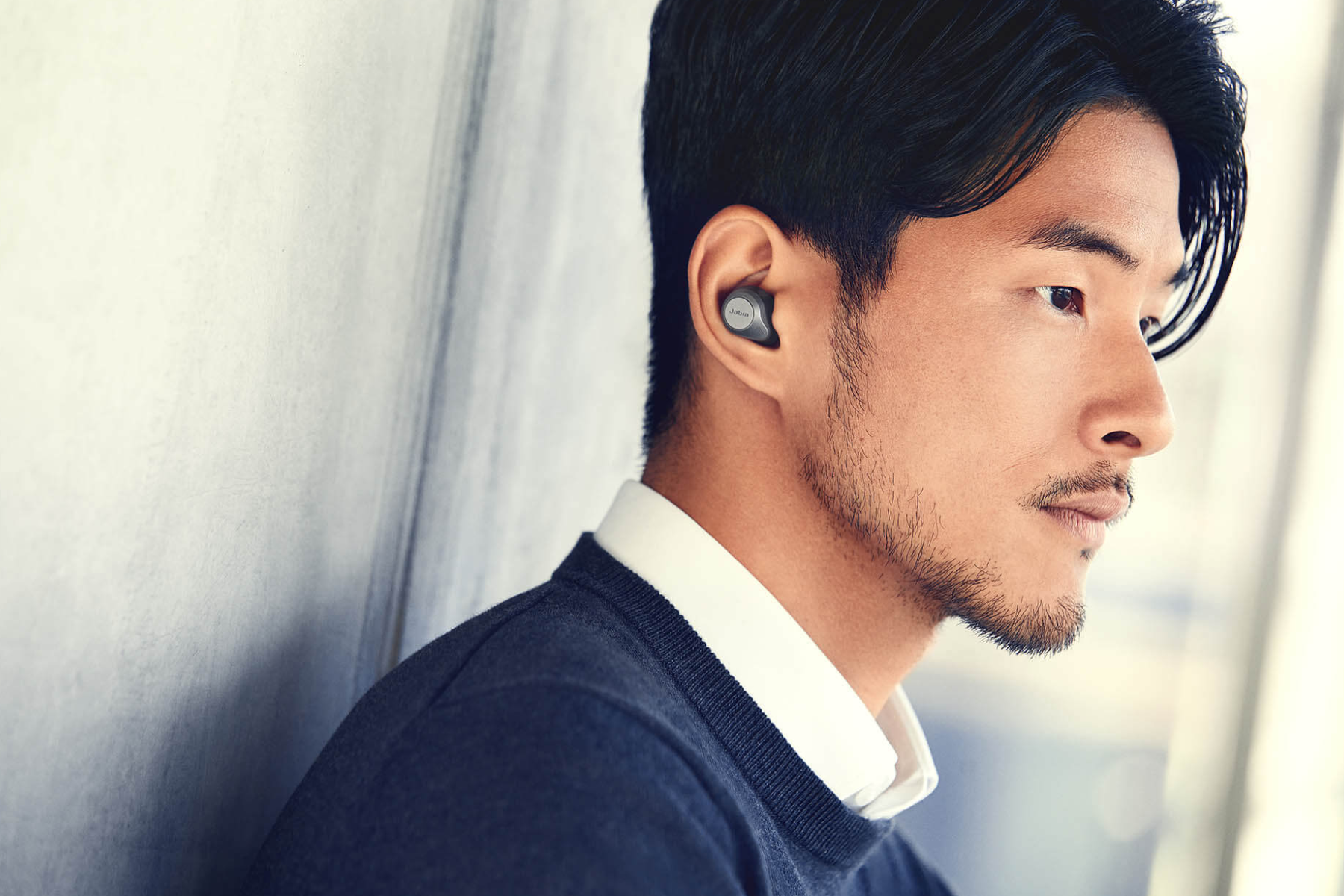 An Asian man is seen leaning on a wall while wearing a pair of Jabra Elite 85t earbuds