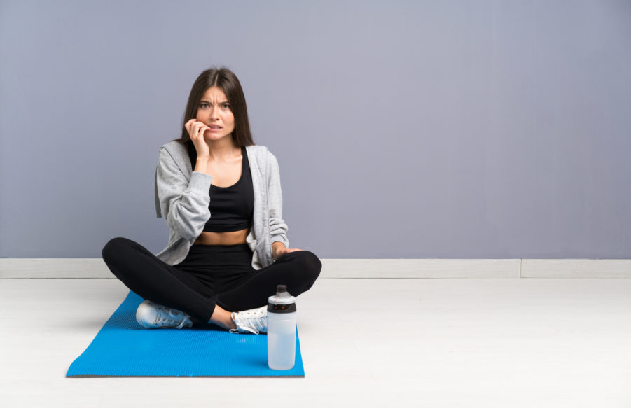A woman in grey hoodie biting her nails while sitting on a yoga mat