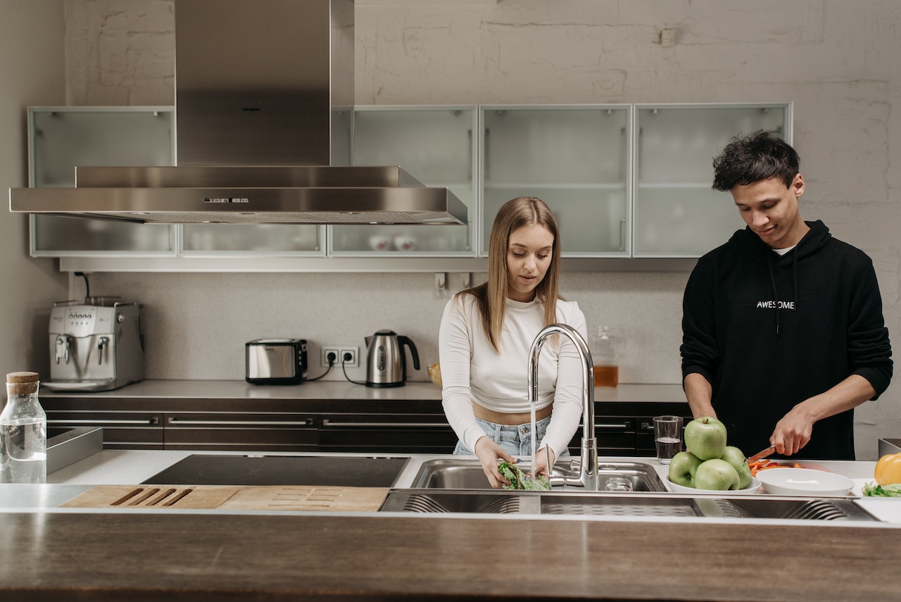 A young couple in the kitchen, with the guy slicing and the girl washing some leafy vegetables