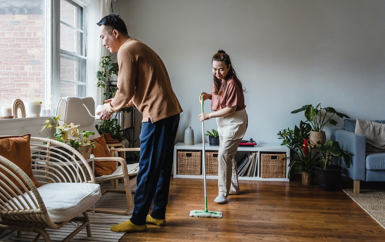 A middle-aged couple in the living room, with the wife mopping and the husband watering the indoor plants