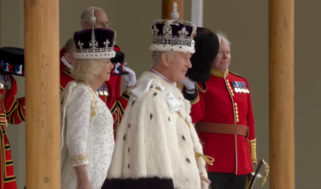 King Charles III and Queen Consort Camilla behind him, both wearing crowns and in their full regalia