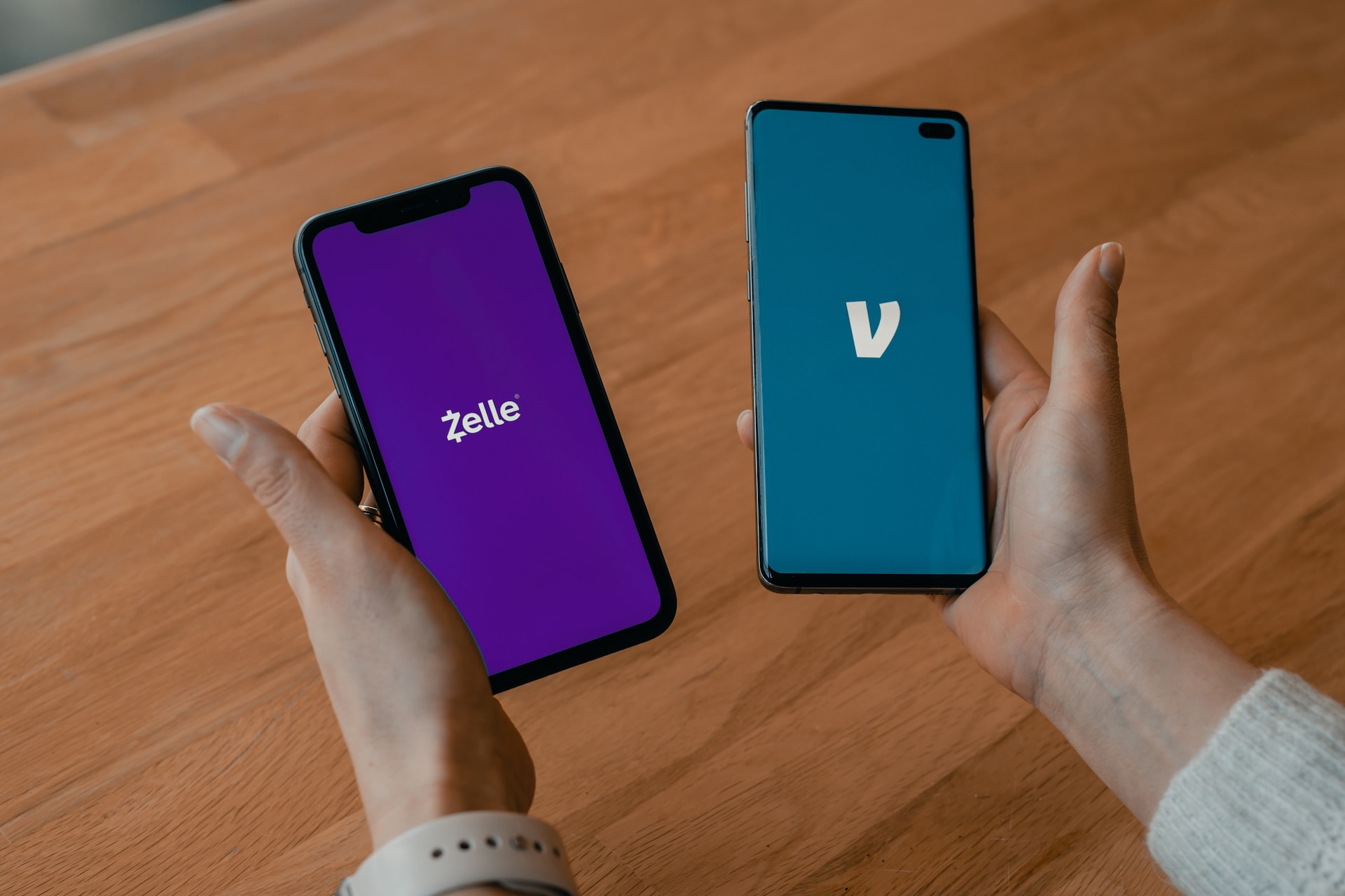 A left hand with a smartphone with ‘Zelle’ on the screen and a right hand with a smartphone with the Venmo ‘V’ logo