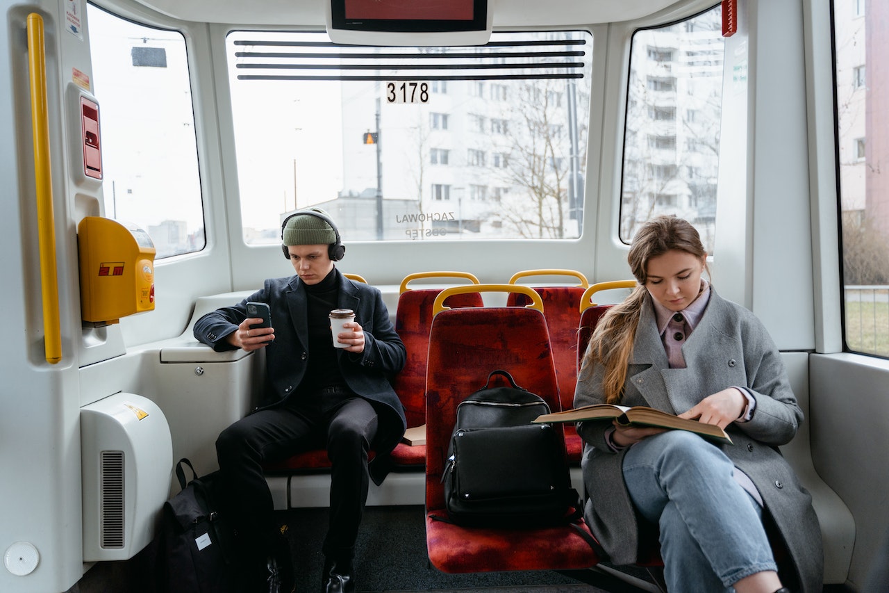 A woman in gray coat reading a book inside a train and sitting near a man wearing headphones and using an iPhone
