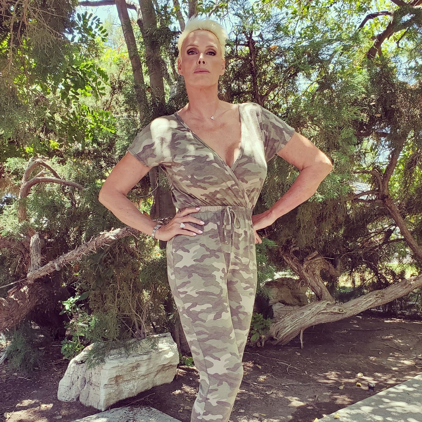 Brigitte Nielsen in camouflaged top and pants, with hands on her hips and some trees at her back