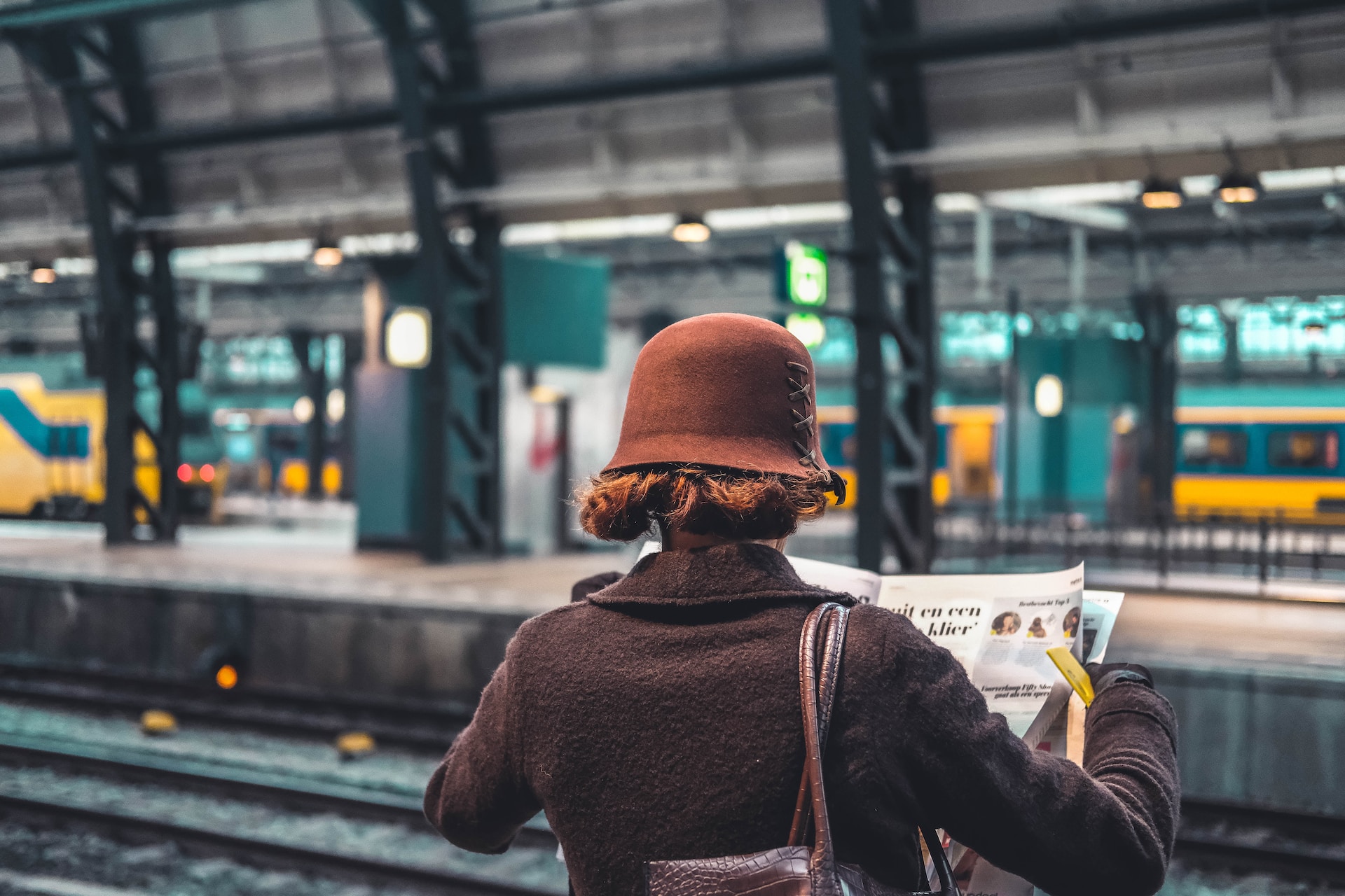 A lady in brown hat and coat standing near the tracks and reading the newspaper in a train station