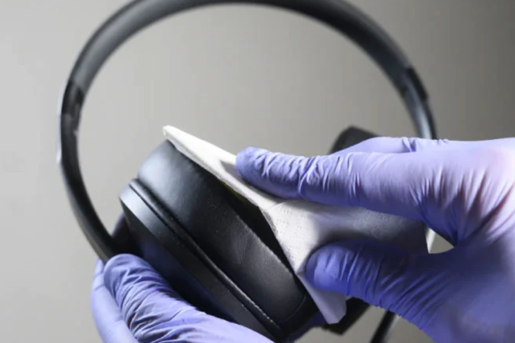 A pair of gloved hands using a tissue to clean a black headphone