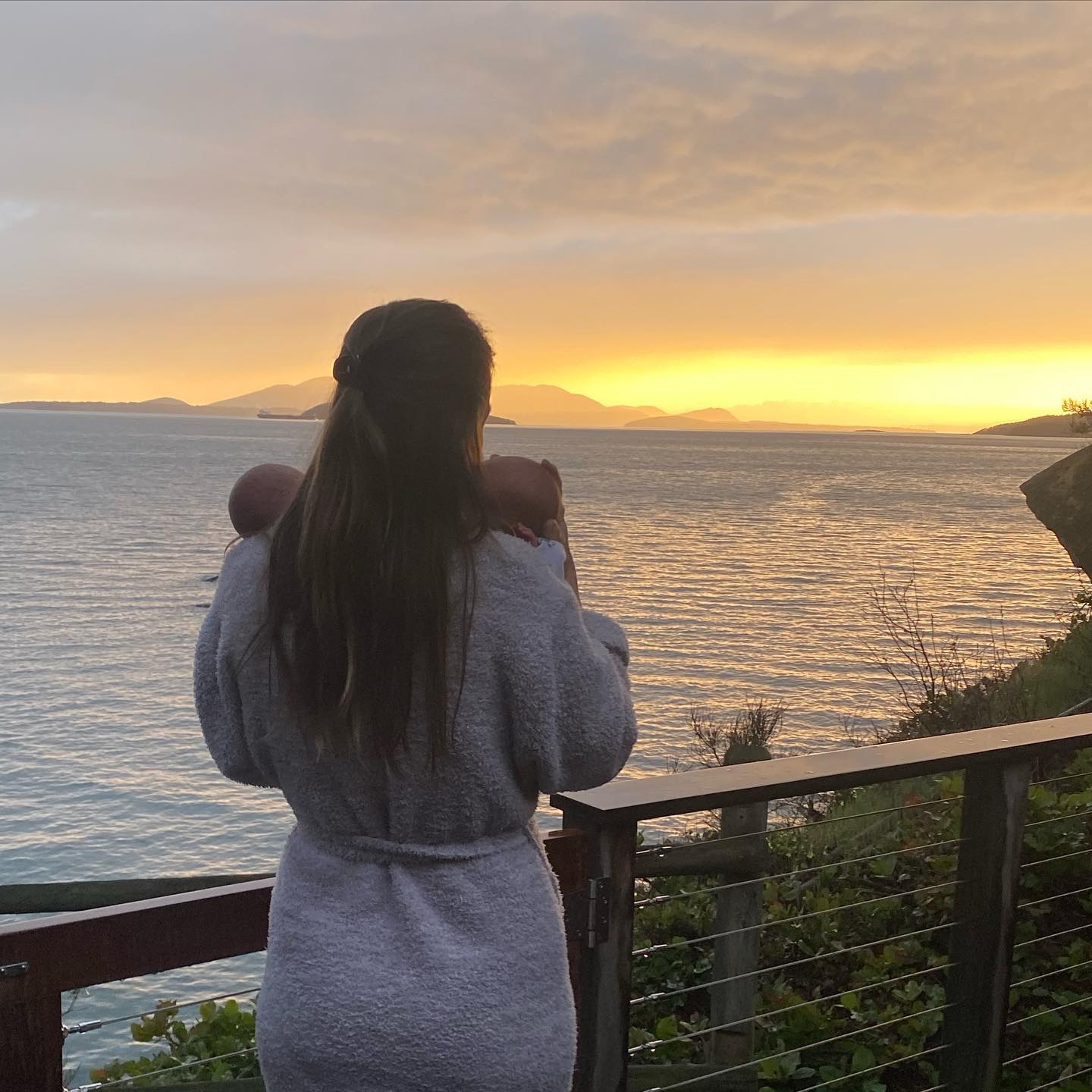 Hilary Swank in a robe holding her twin babies while looking across the water and the sunset