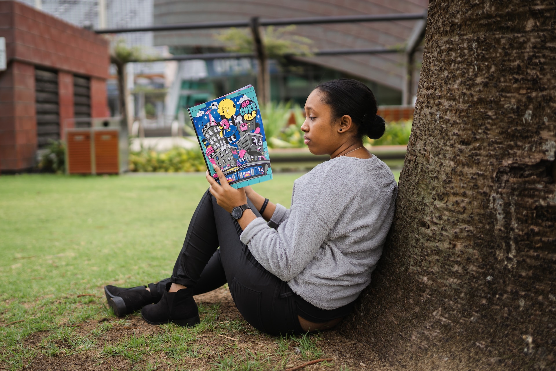 A young Black woman sitting in a park and leaning on a tree while reading No Cure Magazine