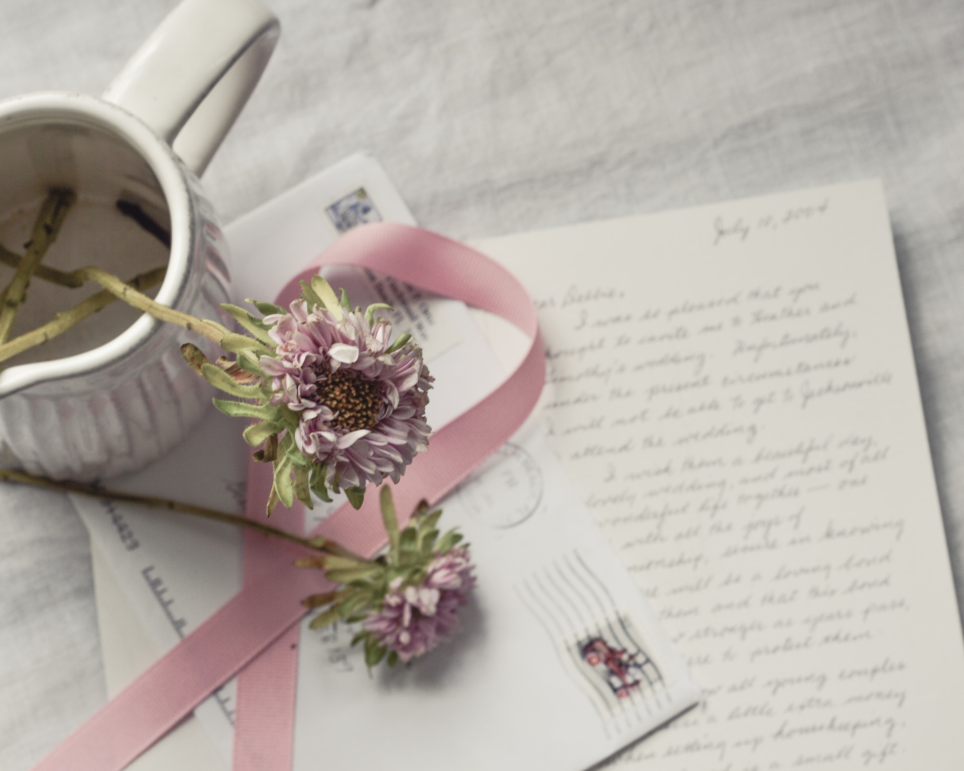 Two flowers, a pink ribbon, a stamped envelope and a handwritten letter on a white paper and in cursive