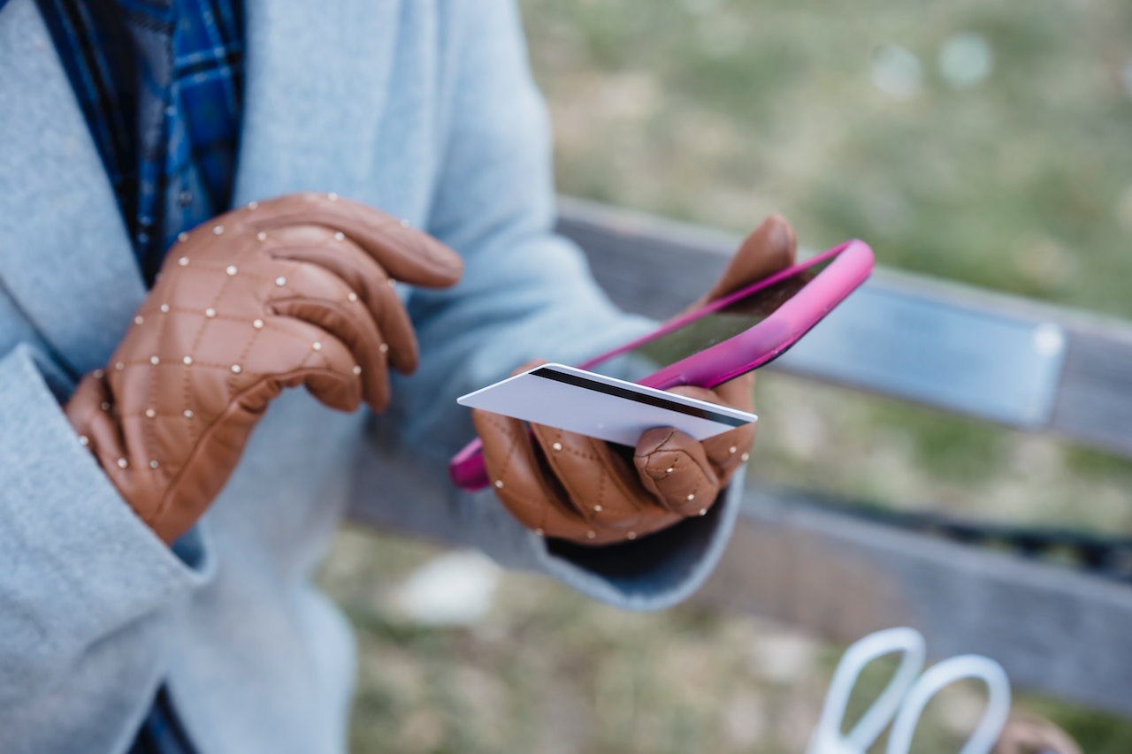 Woman wearing brown leather gloves outdoors and holding a pink smartphone and a credit card