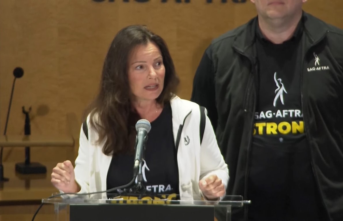 Fran Drescher in black T-shirt under a white jacket speaking behind a podium in a SAG-AFTRA press conference