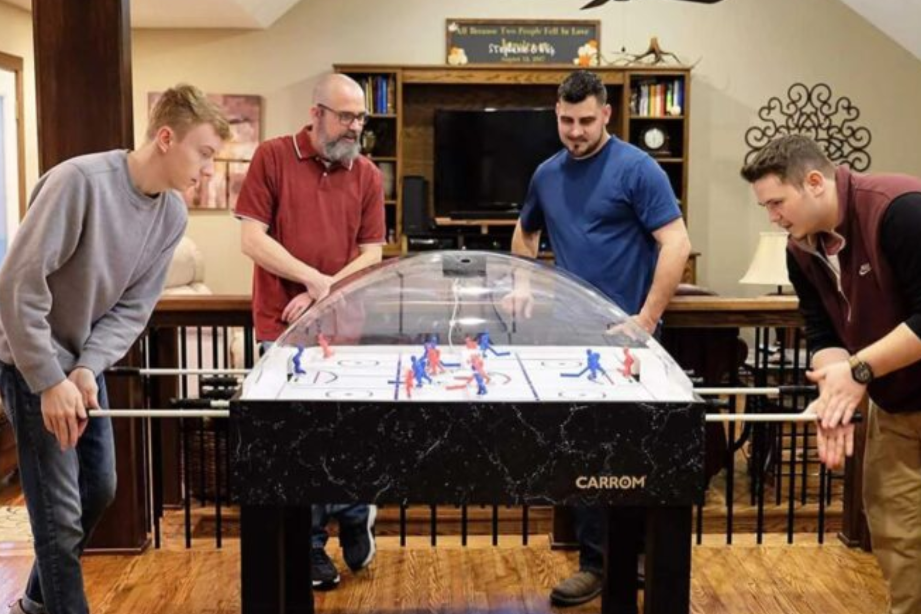 Four individuals gathered around a Bubble Hockey table. Two of them are actively playing the game while the other two are observing the match