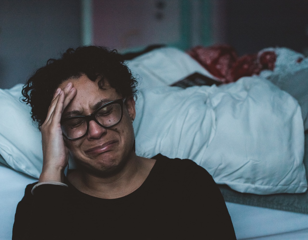 A black woman with glasses sitting on the floor with her back against the bed and crying