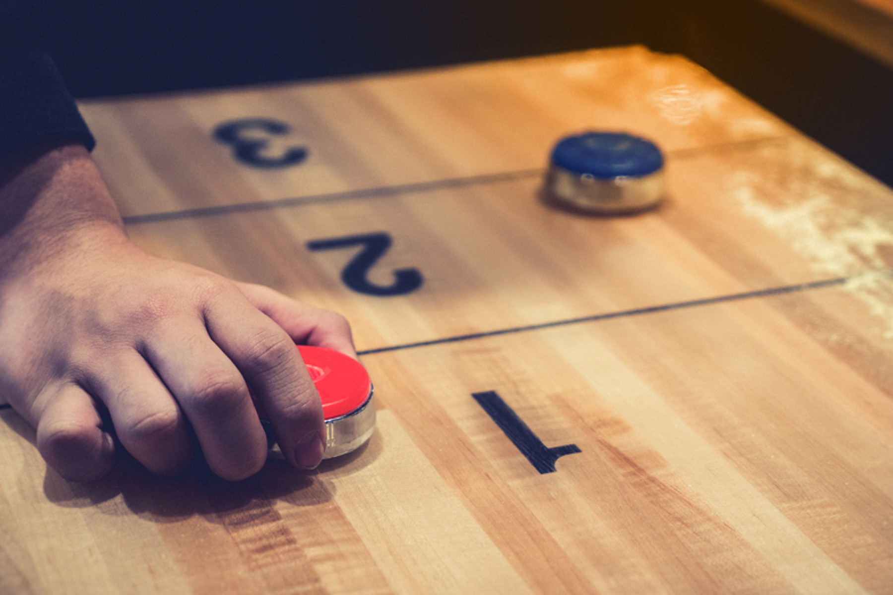 A red puck is being held by a hand next to a blue puck on a shuffleboard