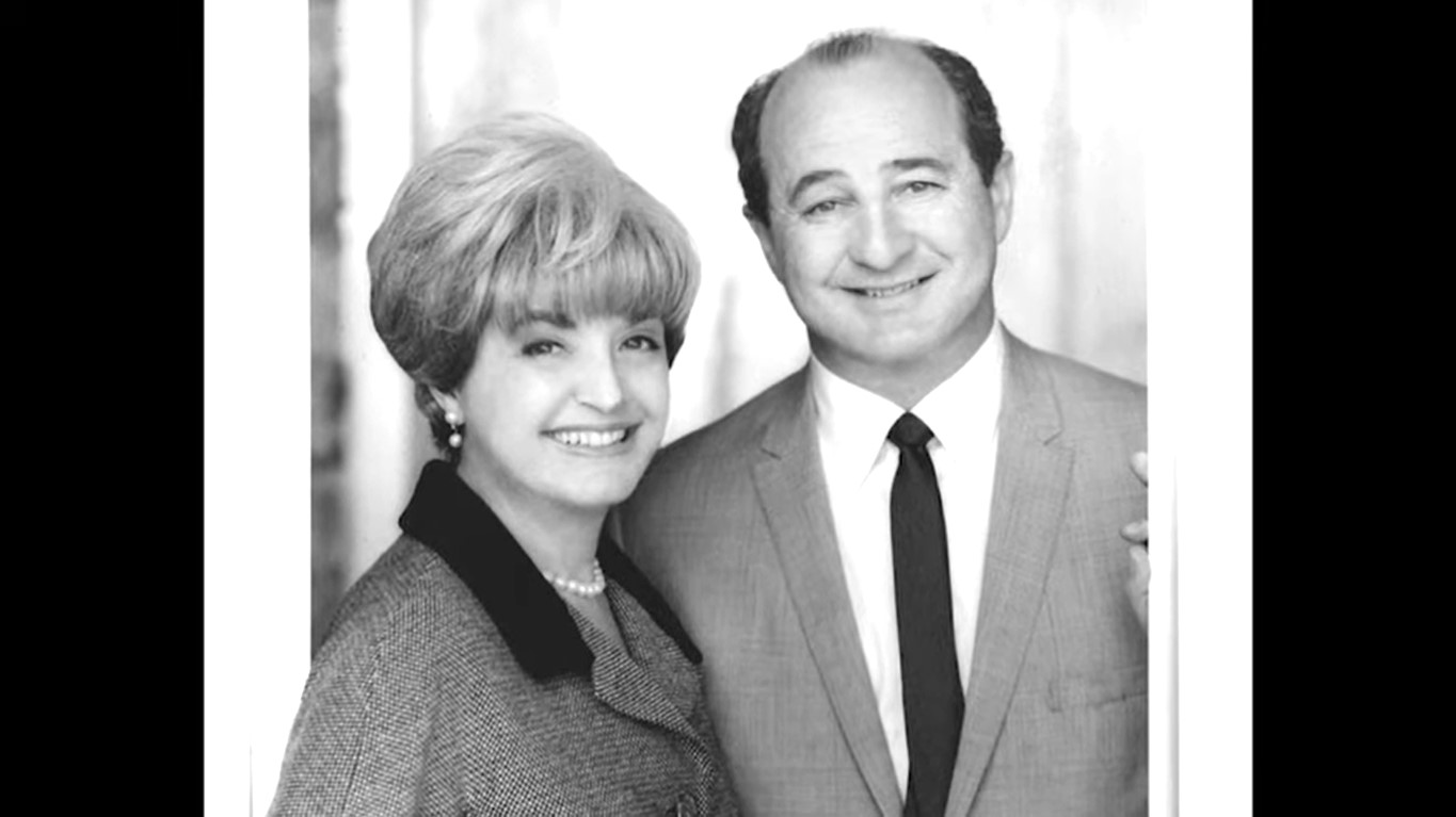 Ruth Handler in pearl necklace and blazer and Elliot Handler in a coat and tie smiling together