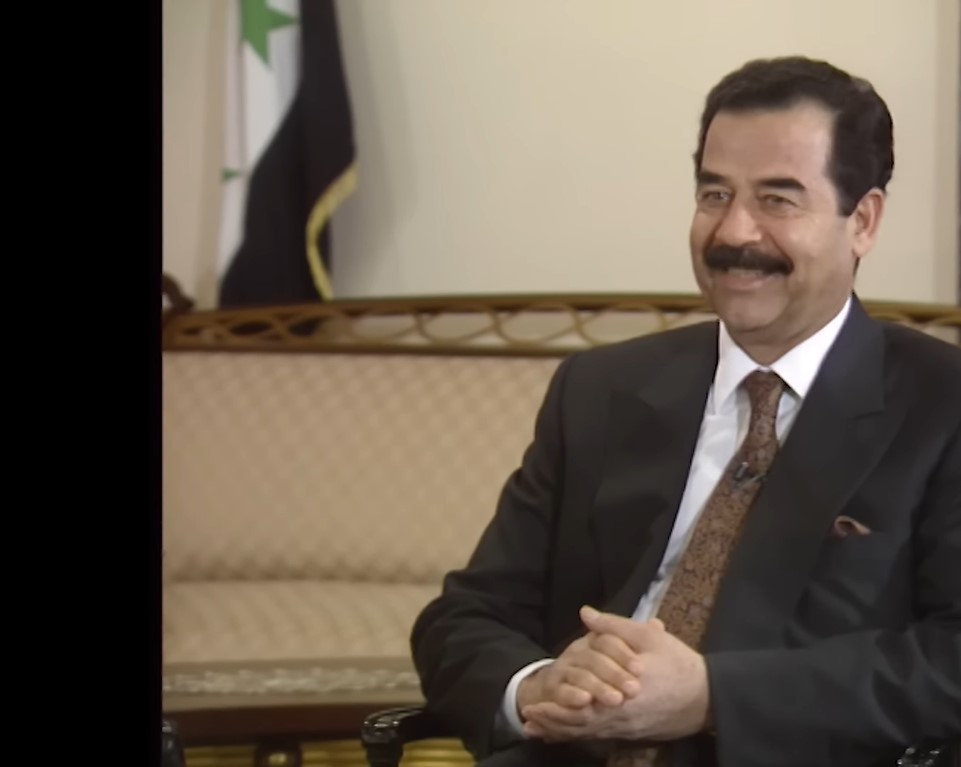 A sitting Saddam Hussein in a dark American suit, smiling and clasping his hands together