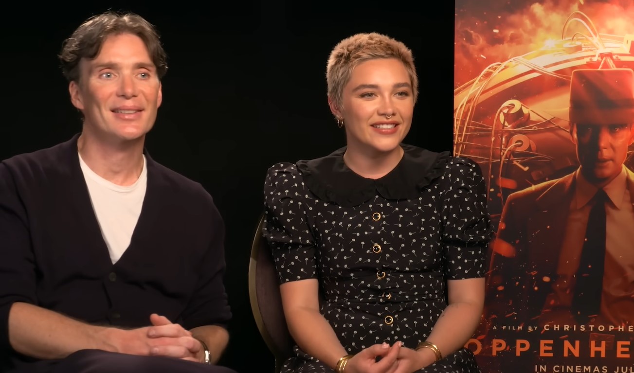A smiling Cillian Murphy and Florence Pugh seated beside each other, with the ‘Oppenheimer’ movie poster