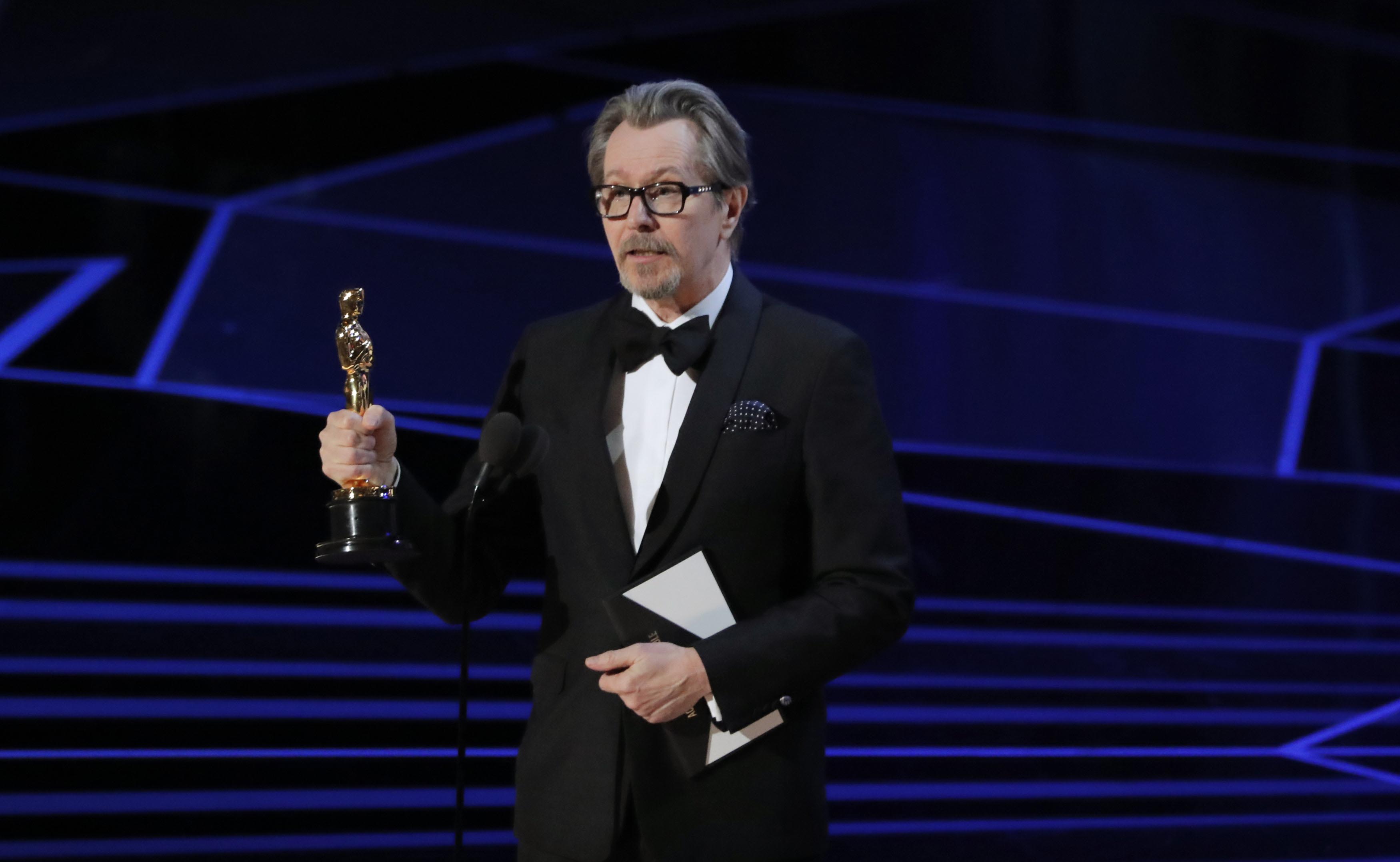 Gary Oldman wearing a black suit while holding an Oscar award trophy