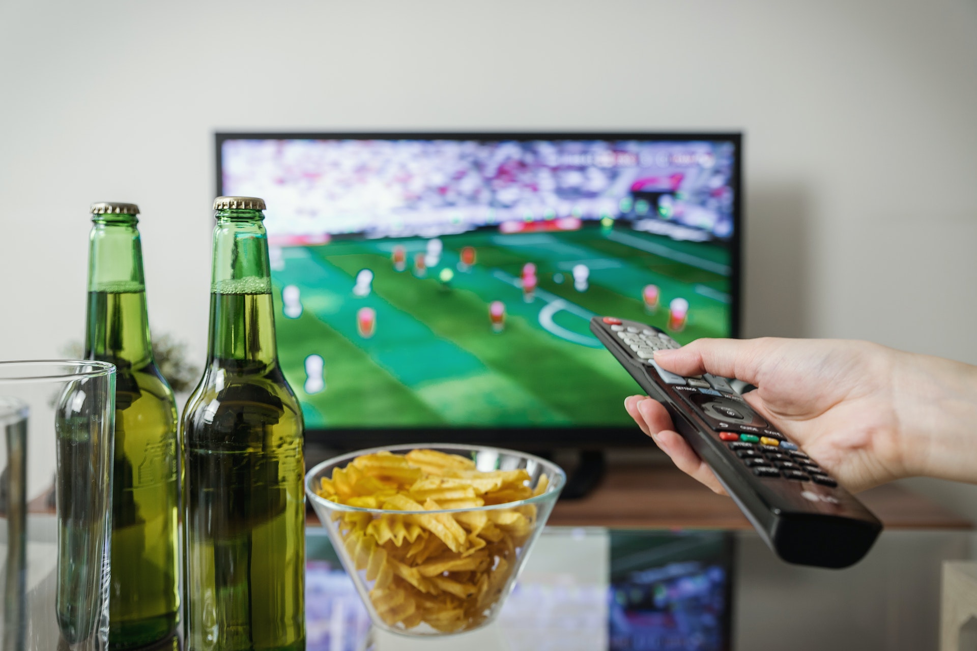 Football match on TV screen while a remote controller in hand and two carbonated drink bottles with glass are also present on table.