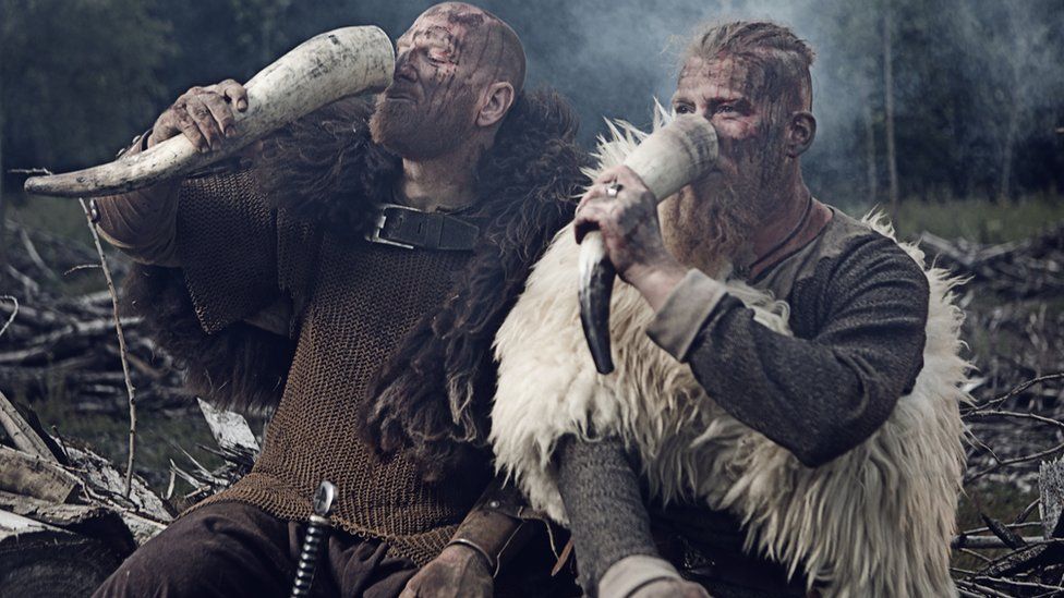 Two Vikings drinking from a skull horn while sitting on a wood log
