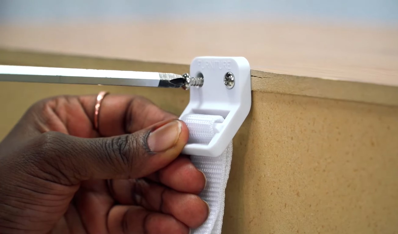 A faceless Black person’s hand screwing a white furniture wall strap on a wooden furniture’s edge