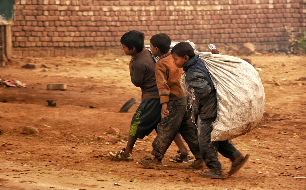 Three poor children carrying a huge sack on their back and walking closely together on a dirt road