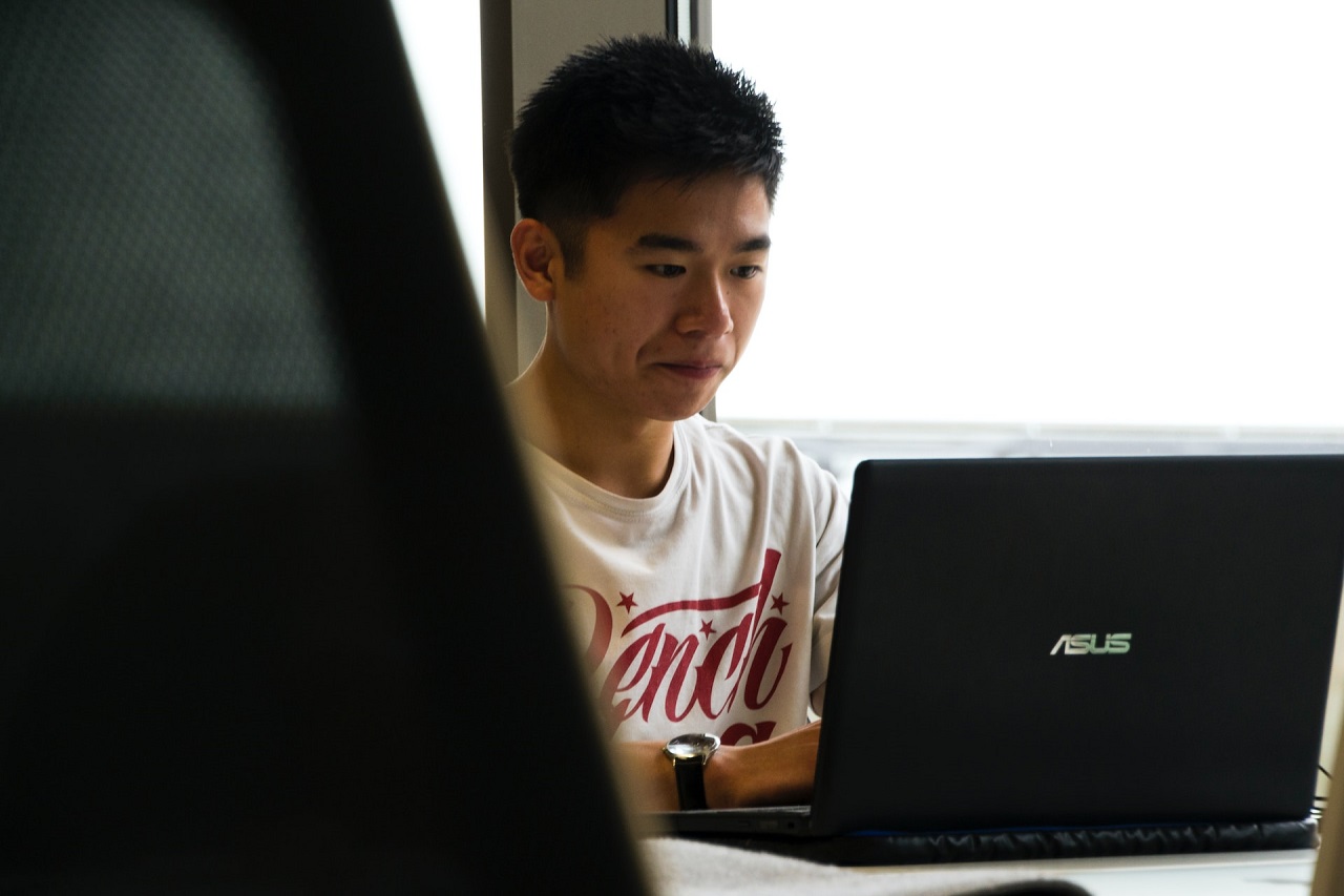 A short-haired Asian male in T-shirt sitting by the window and typing on a black ASUS laptop
