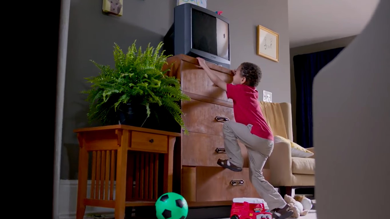 A male Black kid in red polo shirt climbing on a four-layer wooden drawer with a TV on top