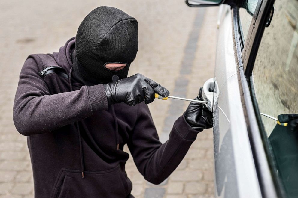 A thief in mask is opening car door with screw driver.