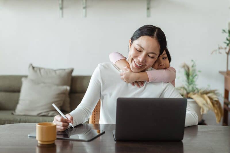 A woman with hers kid is hugging on her back while she is working on laptop and writing.