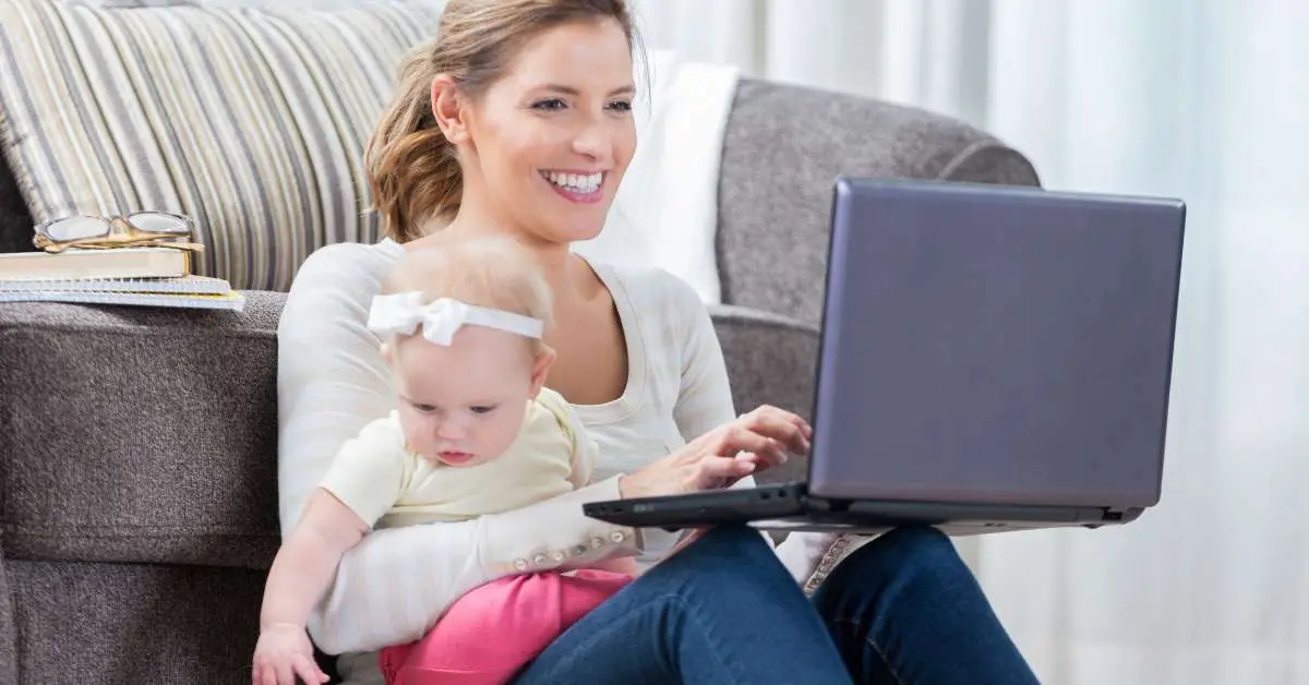 A woman is working on laptop while carrying her child in lap.