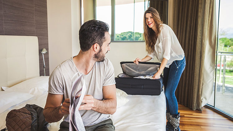 A woman is looking into luggage bag while talking her man who is sitting on bed.