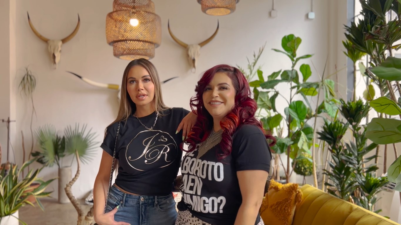 Jenni Rivera and Chiquis posing together in black and tucked-in T-shirts, with indoor plants in the background