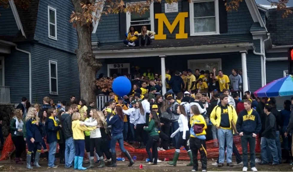 Crowd of people wearing "M" merchandise gather outside a house for an event or celebration.