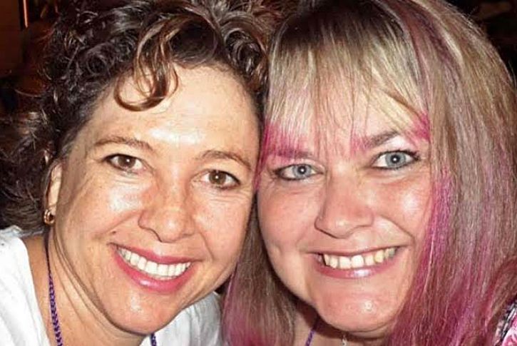 Two women smiling closely for a photo, one with dark, curly hair and the other with straight, blonde hair with pink highlights.
